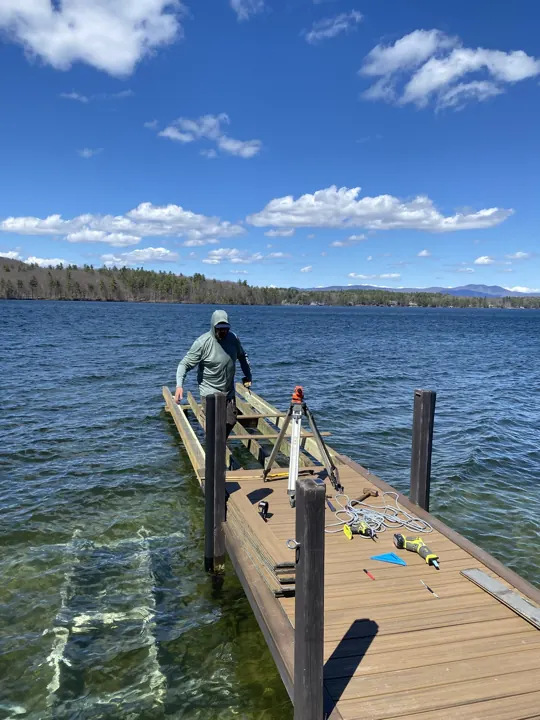 Worker setting dock framing beside a tripod-mounted level over clear lake water