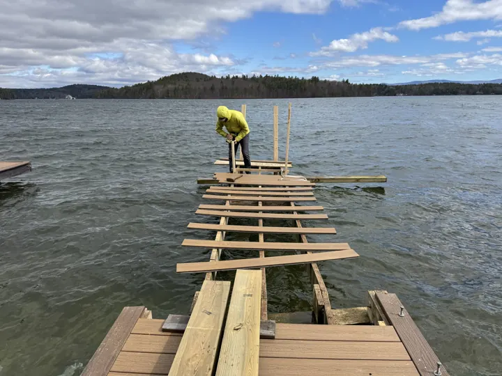 Worker on exposed dock framing, with boards removed and lake water below